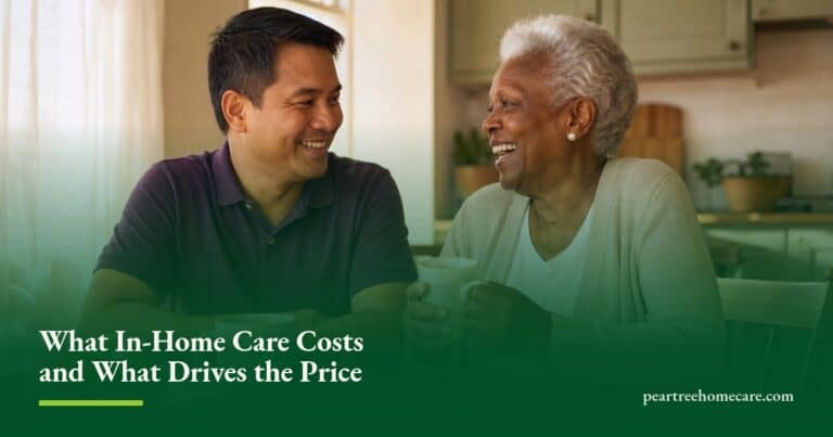A caregiver and an older woman share tea and a laugh at a sunlit kitchen table in her home.